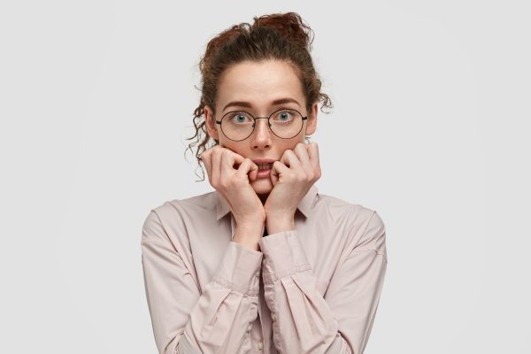 Unhappy nervous young female with worried expression, bites finger nails, looks anxiously directly at camera, wears spectacles, dressed in fashionable clothes, stands against white background.